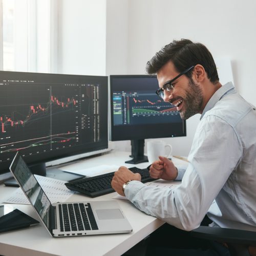 Lucky day. Happy young businessman or trader in formalwear and eyeglasses using laptop and smiling while sitting in his modern office. Stock exchange. Financial trading concept. Investment concept