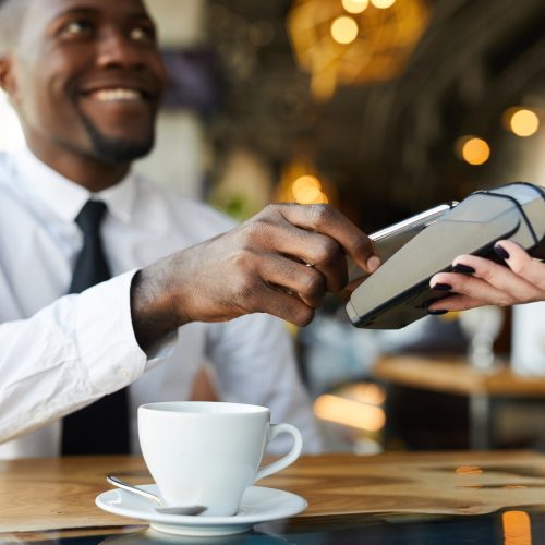 Close-up of positive young African-American businessman sitting at table and using smartphone while making contactless payment in cafe