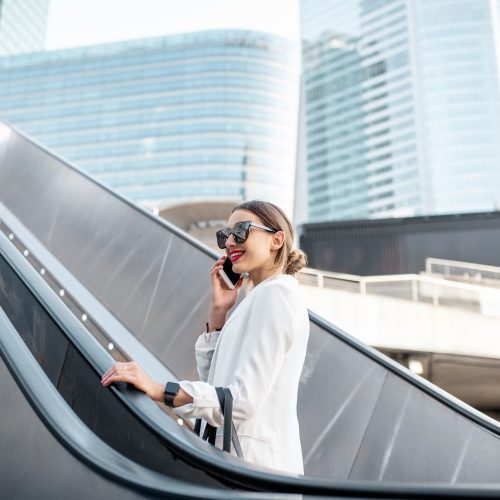 Stylish businesswoman in white suit talking phone while going up on the escalator at the business centre outdoors in Paris